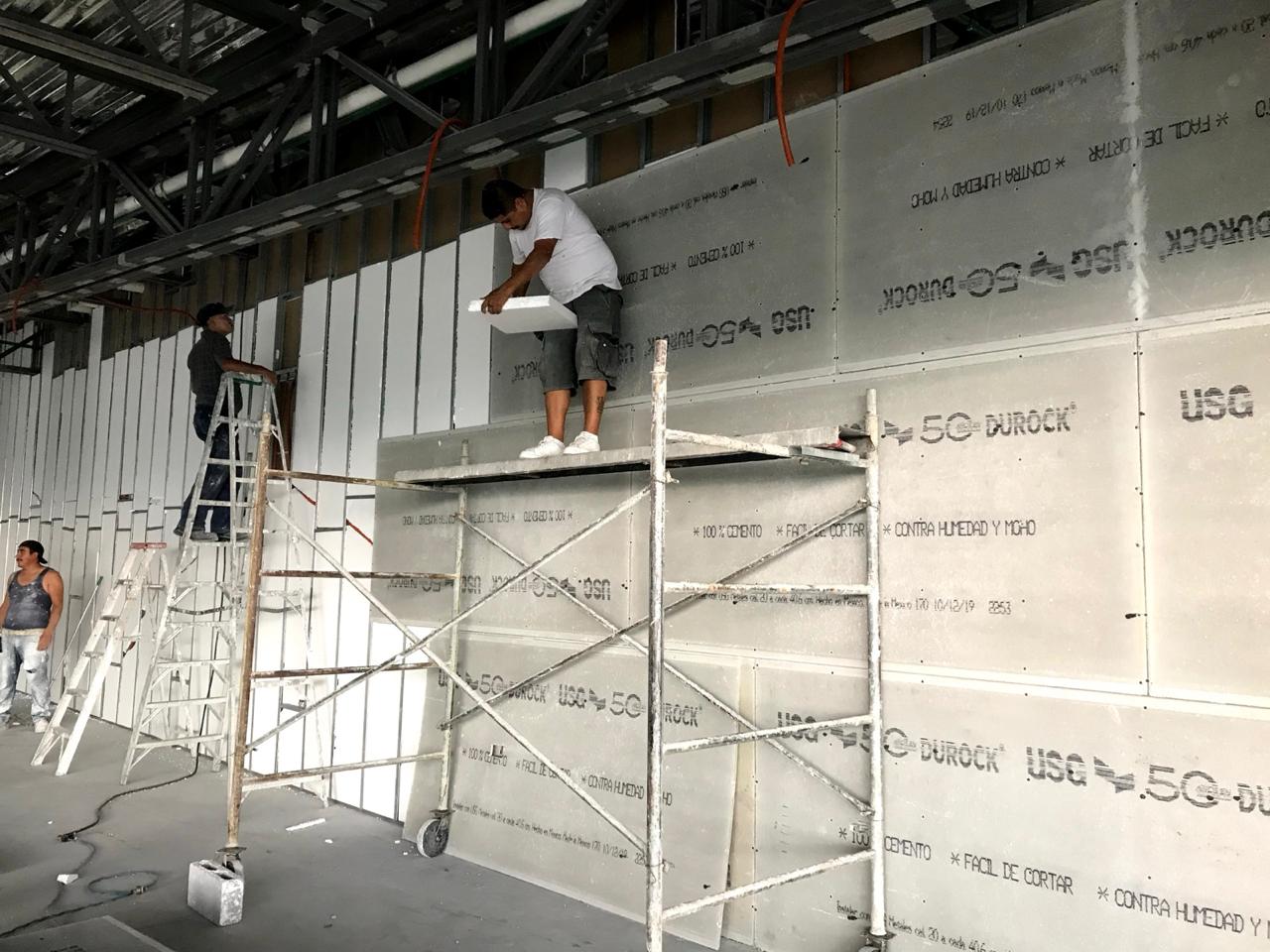 Cuadrilla instalando panel de yeso sobre andamio en planta industrial